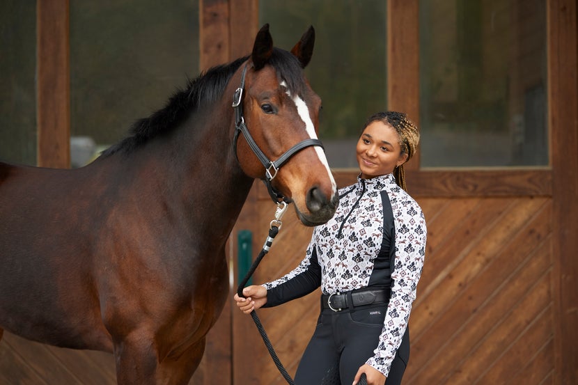 Woman wearing the Kerrits Always Cool Long Sleeve Shirt in Tan Painted Horse Print, standing next to her haltered bay horse with a white blaze. 