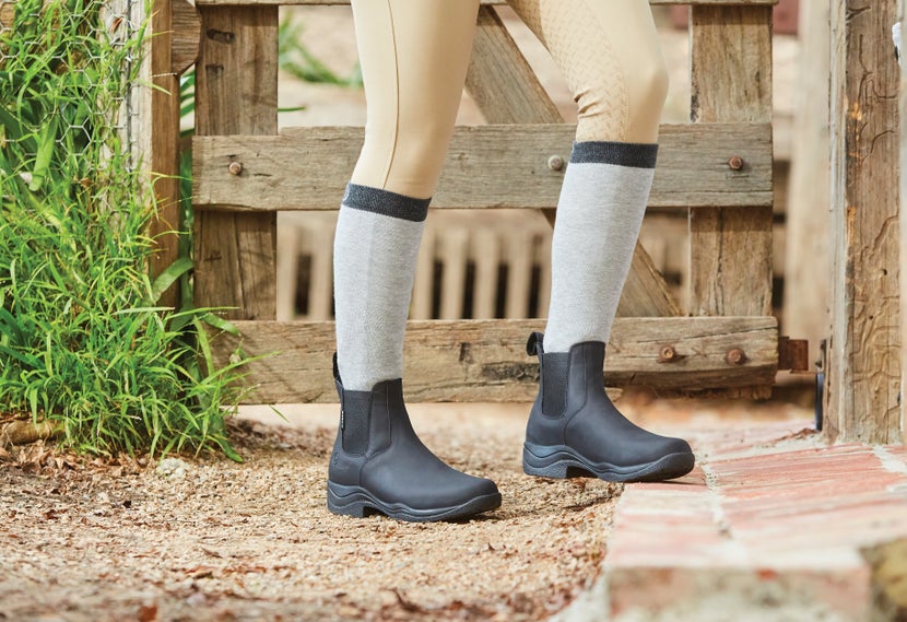 The black Dublin Venturer III Pull-On Riding Boots on someone wearing grey knee-high socks over tan breeches. 