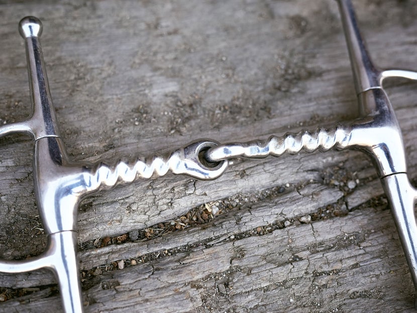 Close up image of a horse bit with a corkscrew mouthpiece, laying flat on a piece of wood. 