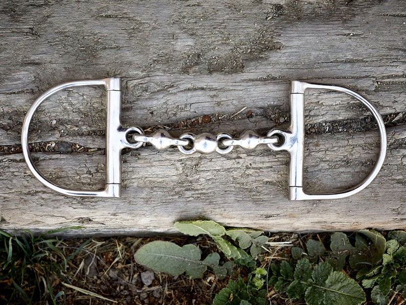 A horse bit with D-ring cheekpieces, laying flat on a piece of wood. 