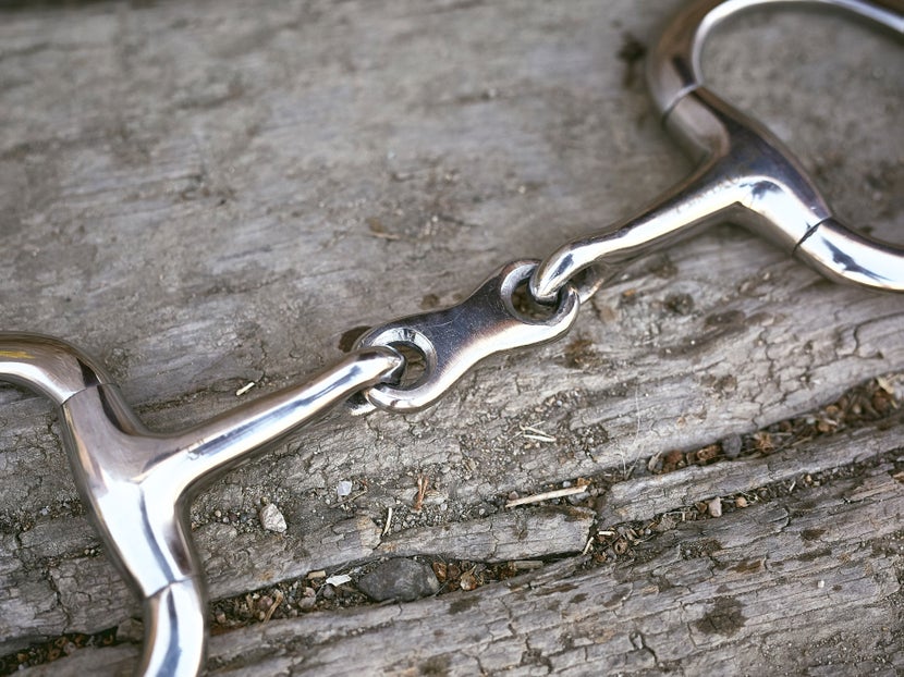 Close up image of a horse bit with a french link mouthpiece, laying flat on a piece of wood. 