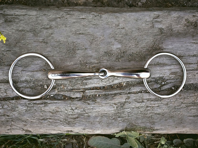 Close up image of a horse bit with a hollow mouthpiece, laying flat on a piece of wood. 