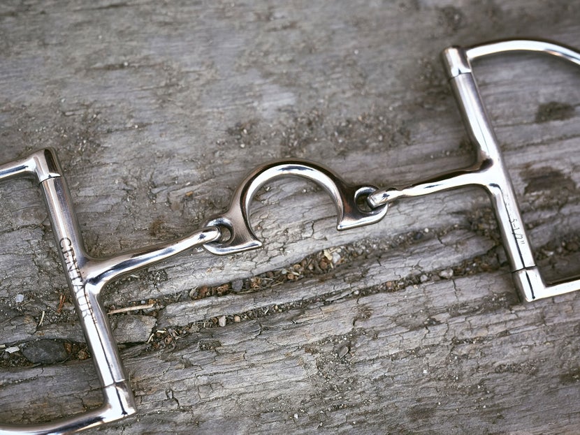 Close up image of a horse bit with a jointed port mouthpiece, laying flat on a piece of wood. 