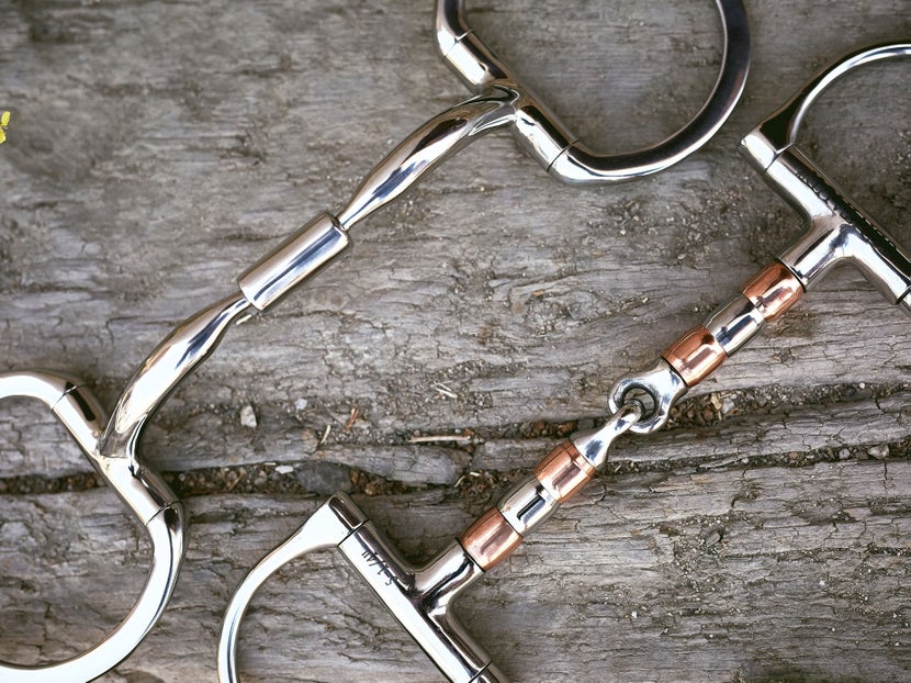 Close up image of two horse bits side-by-side with a roller mouthpieces, laying flat on a piece of wood. 