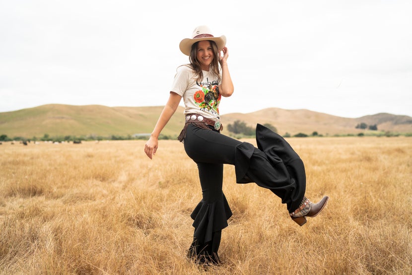 Girl in a field wearing a cowboy hat, vaquero style pants, and fashion cowboy boots.