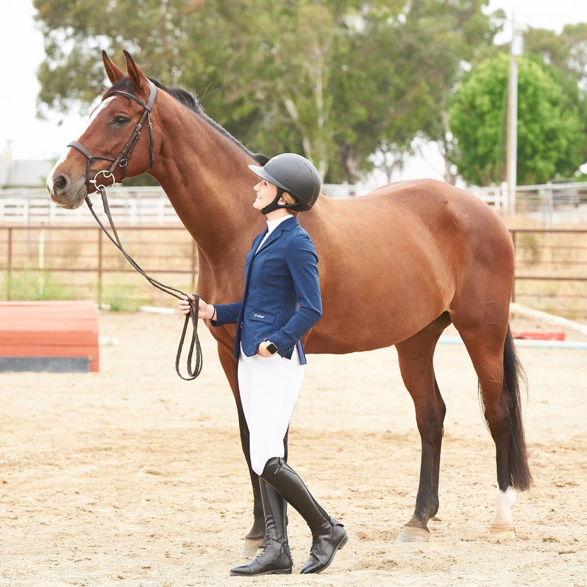 girl wearing an blue EGO7 show coat and tall boots, standing next to her bay horse.