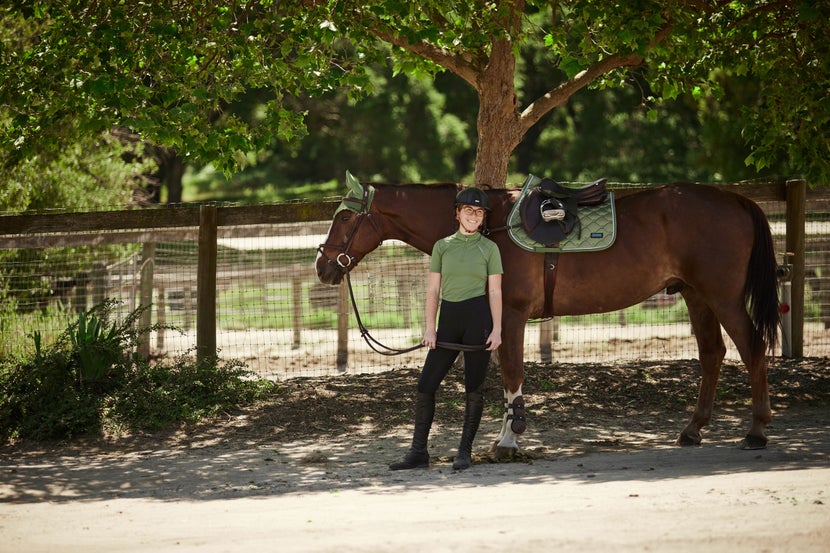Woman standing next to her tacked up bay horse, wearing matching Cavallo tack and riding apparel in green. 