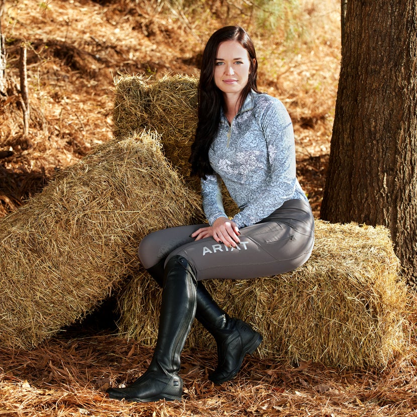 A woman sitting on a hay bale next to a tree, wearing the Ariat V Sport Tall Zip Boots.