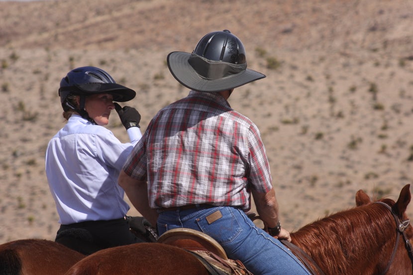 Two horseback riders in the desert, with sun visors on their helmets. 