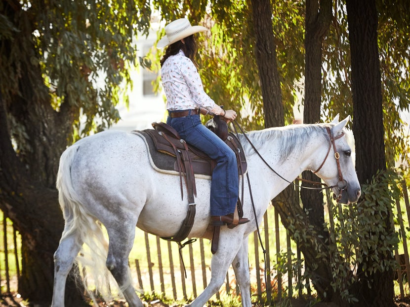 A rider wearing a straw cowboy hat, patterned button down shirt, and dark jeans on her white horse in western tack.