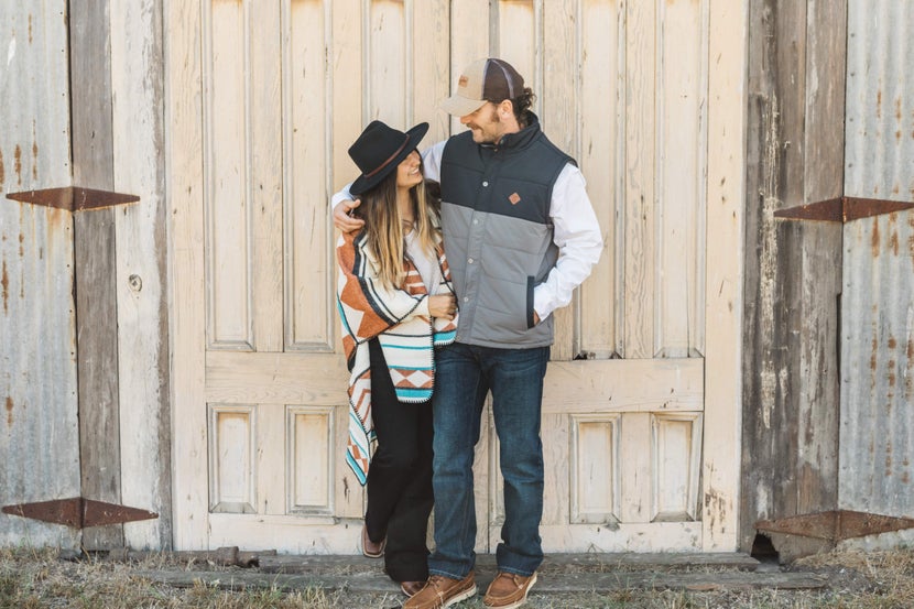 A man and a woman dressed in dark-wash denim, in slightly more formal Western clothes in front of a wooden door.