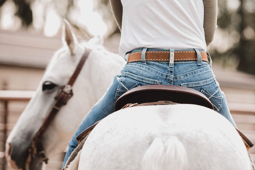View from behind: a rider in a western saddle cut off just above the belt on a white horse, with the horse turning its head so you can see its eye.
