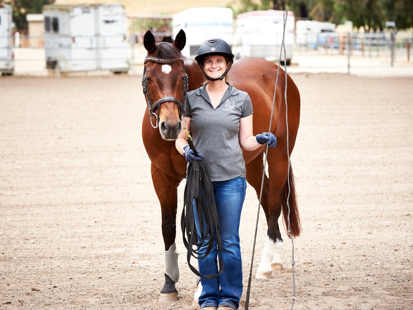 A woman leads her horse with lunging equipment. 