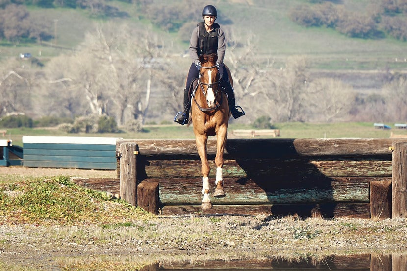 Bec riding a chestnut horse over a wooden jump on a cross country course. 