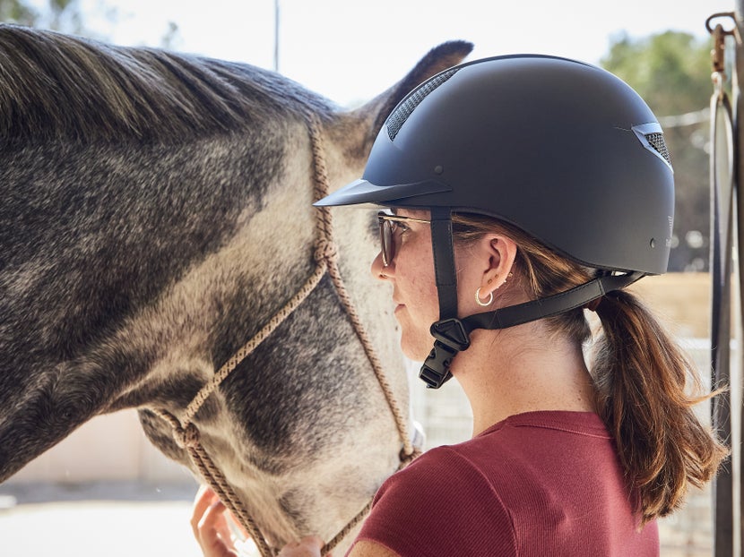 A woman with a secured helmet, standing next to her haltered grey horse.