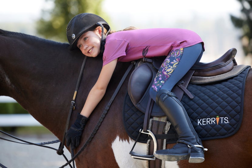 Young girl on horseback leaning over the neck of a paint horse, wearing Kerrits tights and a purple top.