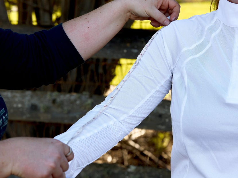 A rider dressed in a white show shirt and their buddy helping take their sleeve length measurement with a soft measuring tape. 