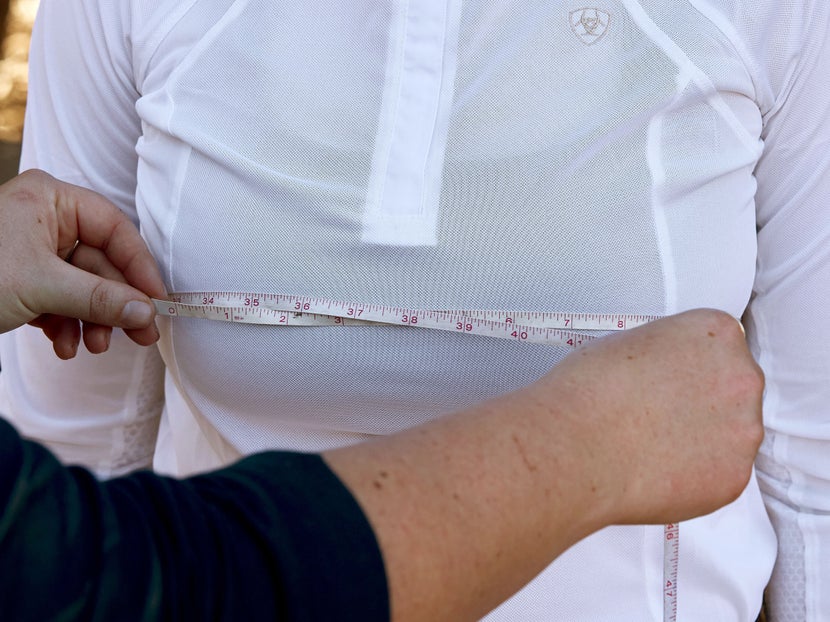 A rider dressed in a white show shirt and their barn buddy helping measure their chest with a soft measuring tape.
