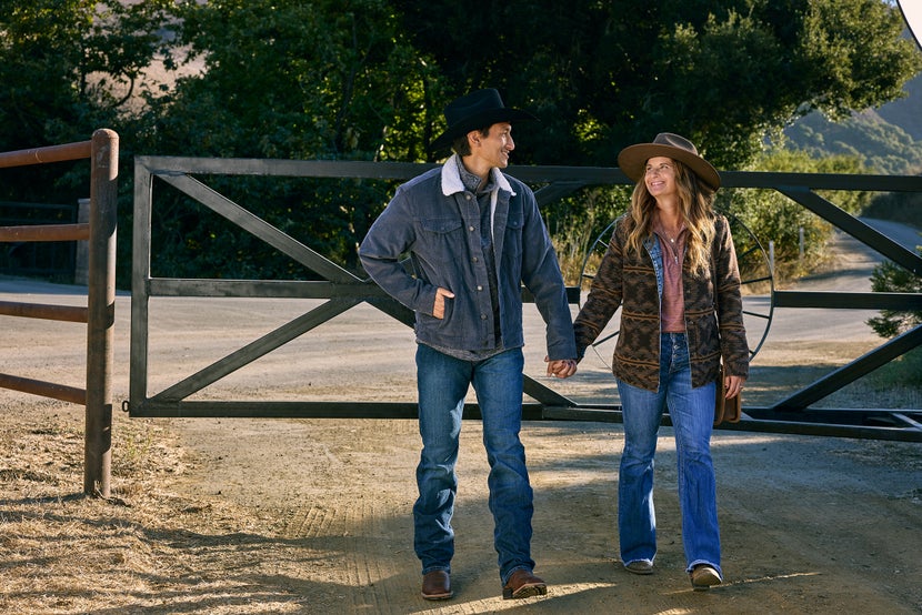 Man and woman walking down dirt path in front of gate hand-in-hand while wearing Western attire, jackets, and cowboy hats. 