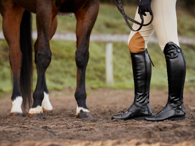 A woman standing next to her horse in an arena, wearing the Ovation Sofia Tall Field Boots.