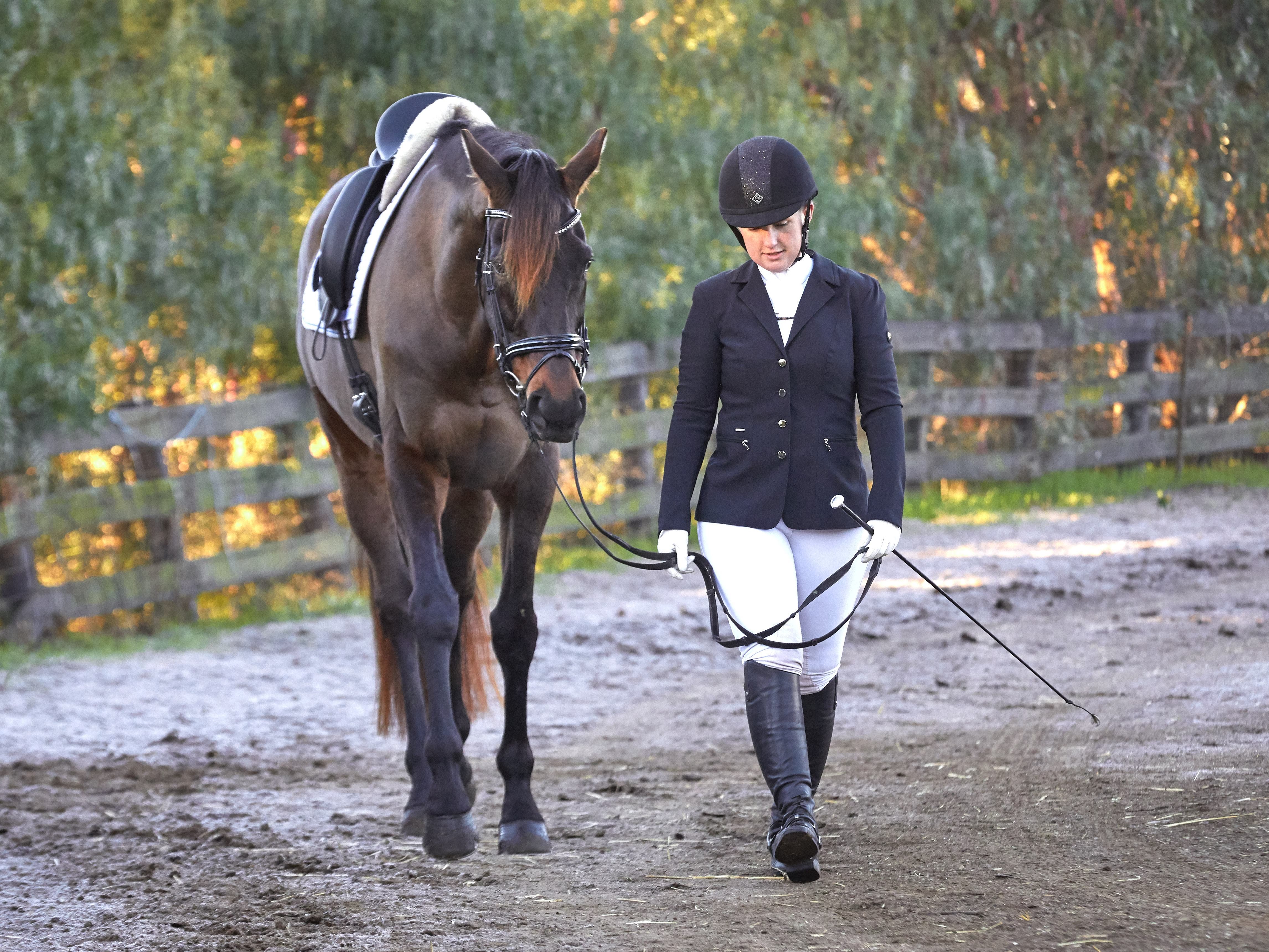 A women walking next to her bay horse, wearing a black button down dressage coat.