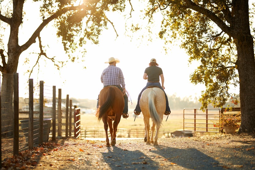 Two riders on horseback at sunset, riding along a fenceline underneath trees.