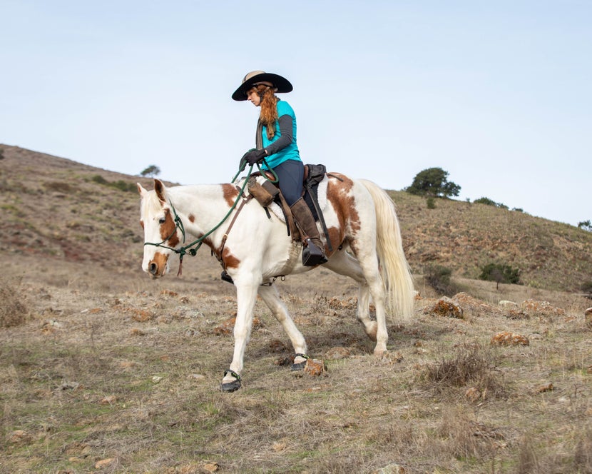 A trail rider riding her pinto horse across rocky terrain, with Explora Magic hoof boots on. 
