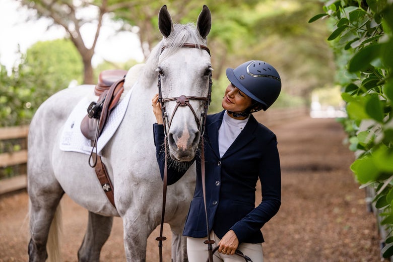 Woman wearing a Charles Owen Kylo Navy helmet, navy show coat, and tan breeches, standing with her grey horse wearing brown tack.