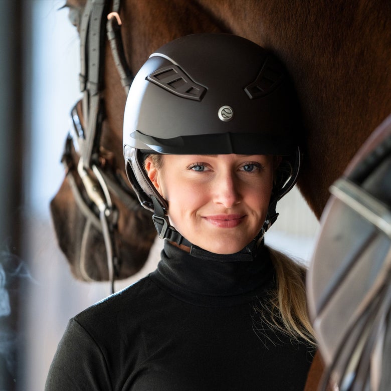 Woman wearing a Trauma Void Lynx helmet and long sleeve black top, standing next to a bay horse in the background.