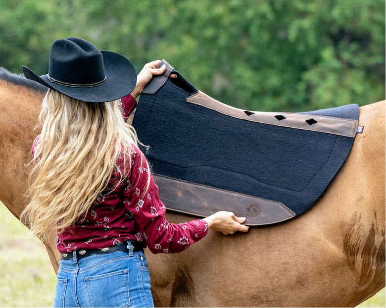 Lady with long blonde hair wearing a black cowboy hat, red floral top, and blue jeans putting the Total Saddle Fit Felt PLUSH Western Saddle Pad on her horse's back.