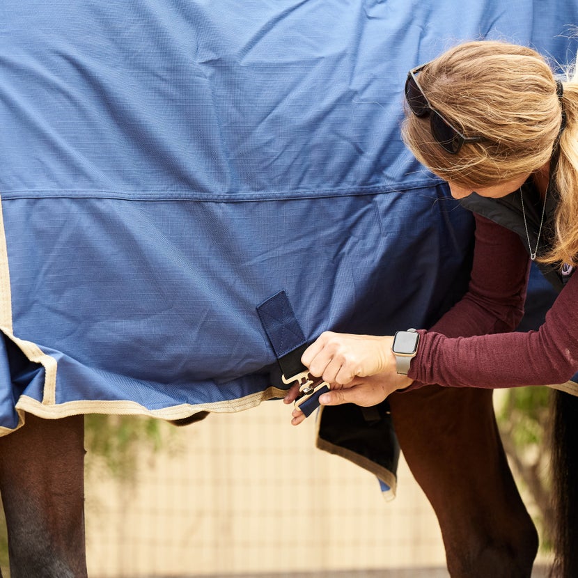 Girl attaching a belly surcingle on a blanket. She is facing toward the horse's head so she's safe if the horse kicks out.